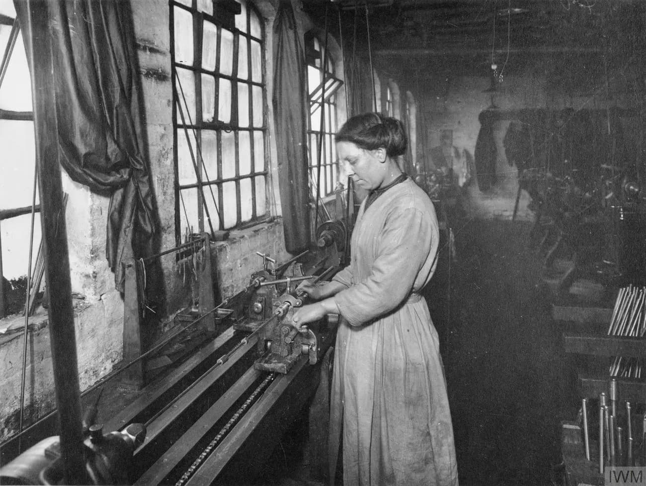 A woman in a long dress operates machinery in a factory, with large windows visible in the background.